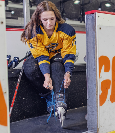 A teenage girl dressed in a hockey uniform laces up her ice skates.